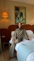 Woman seated on a bed wearing the NaSignature taupe 2-piece set with a brown hijab